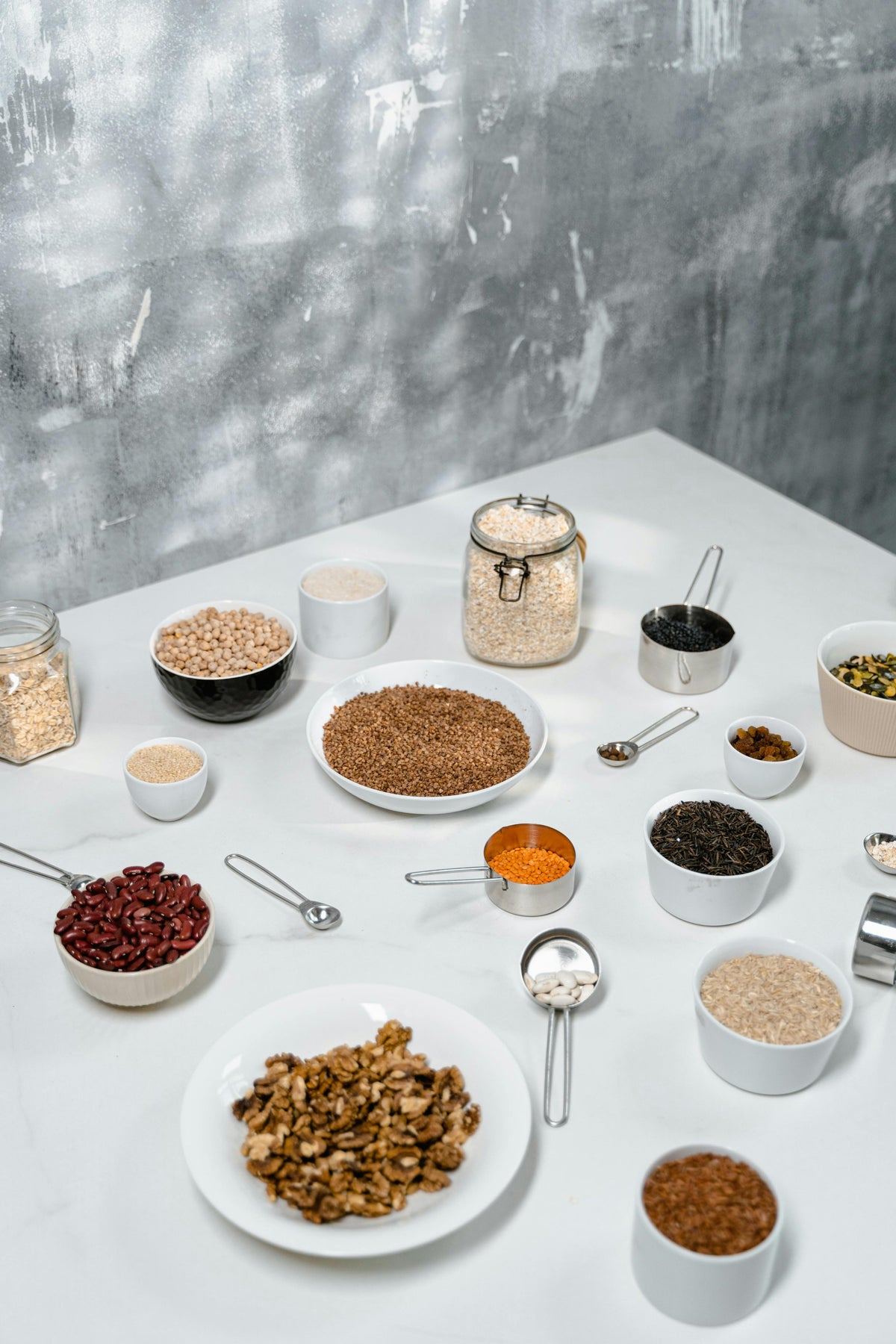 Bowls and measuring cups of legumes, nuts, and whole grains arranged on a white tabletop.