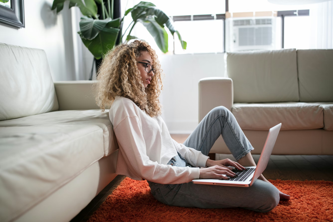 Young woman with curly hair and glasses sitting on the floor against a sofa, typing on a laptop on an orange rug.