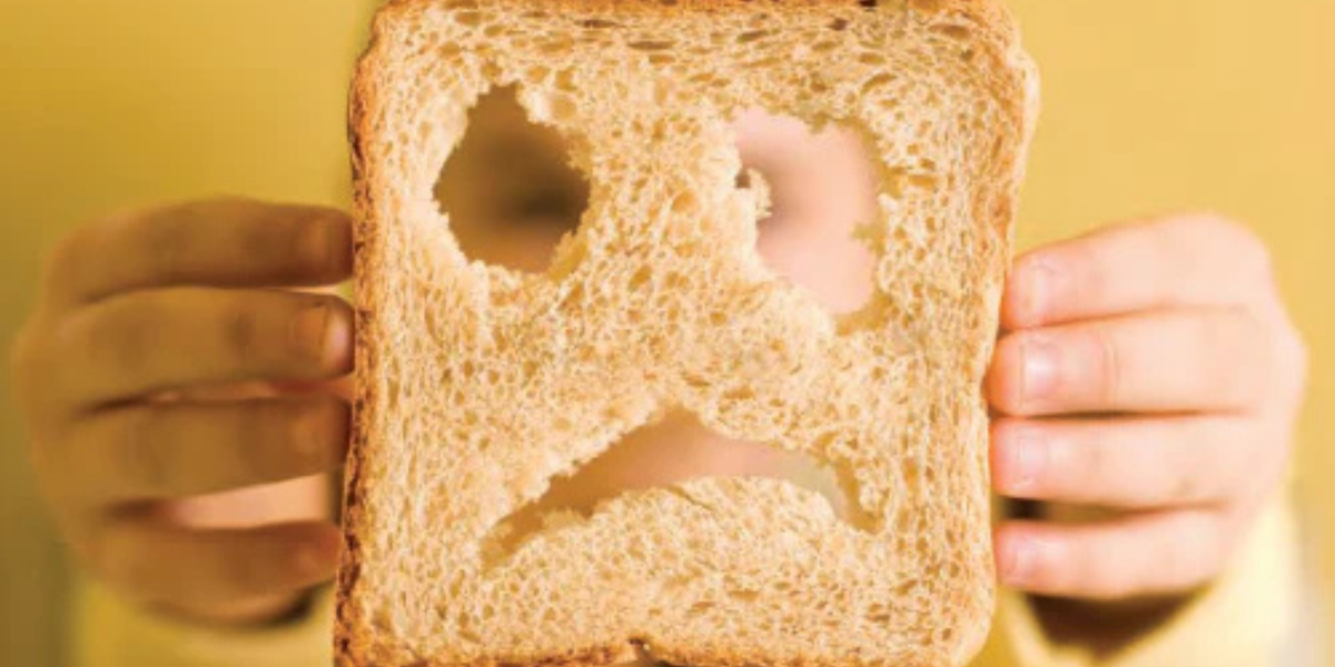 Slice of whole-wheat bread with holes forming a sad face, held by a child's hands.