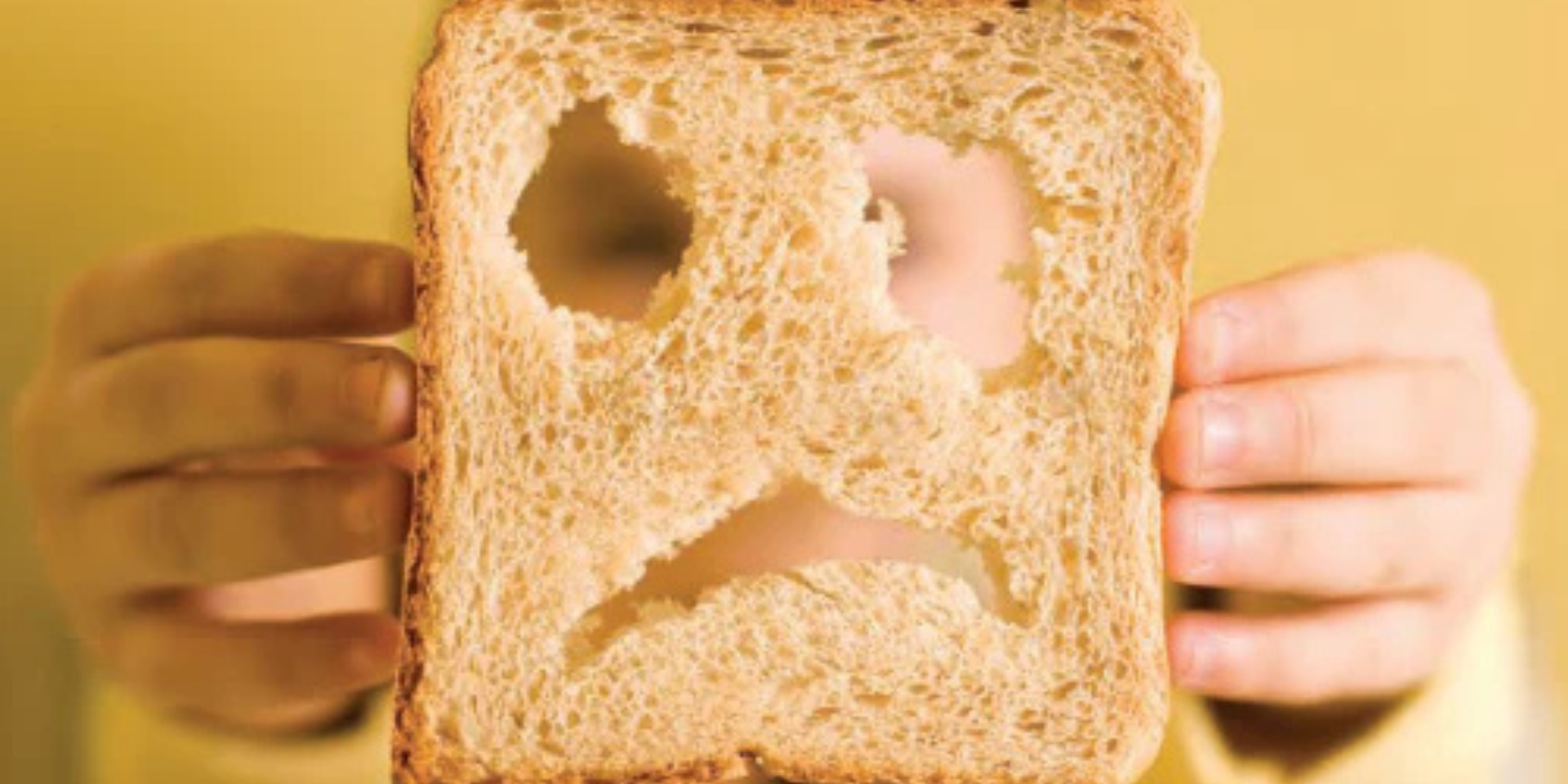 Slice of whole-wheat bread with holes forming a sad face, held by a child's hands.