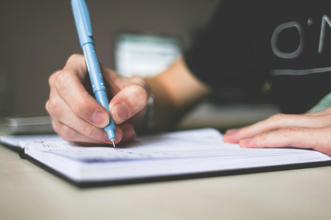 Close-up of a person writing in a notebook with a blue pen on a table, shallow depth of field