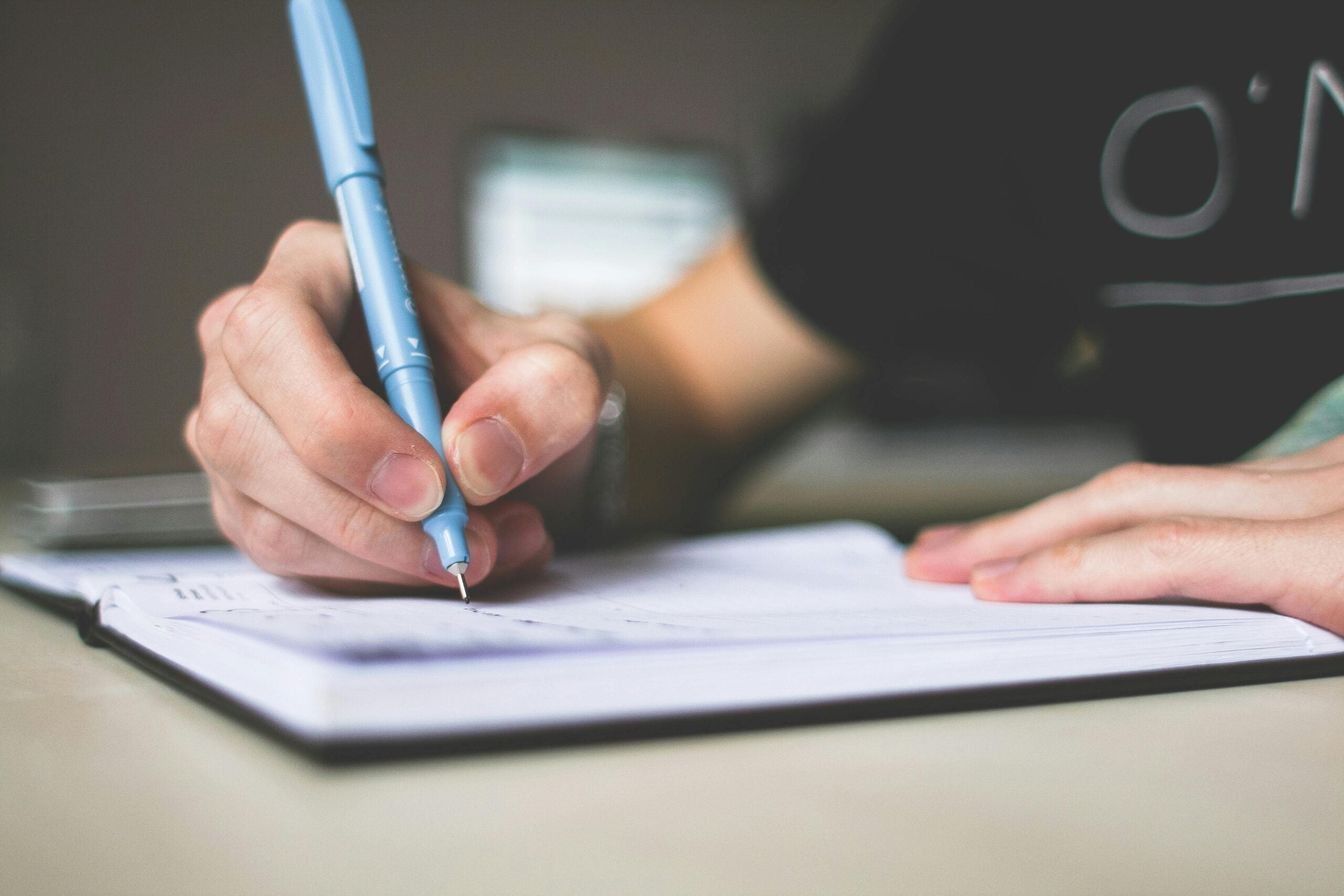 Close-up of a person writing in a notebook with a blue pen on a table, shallow depth of field