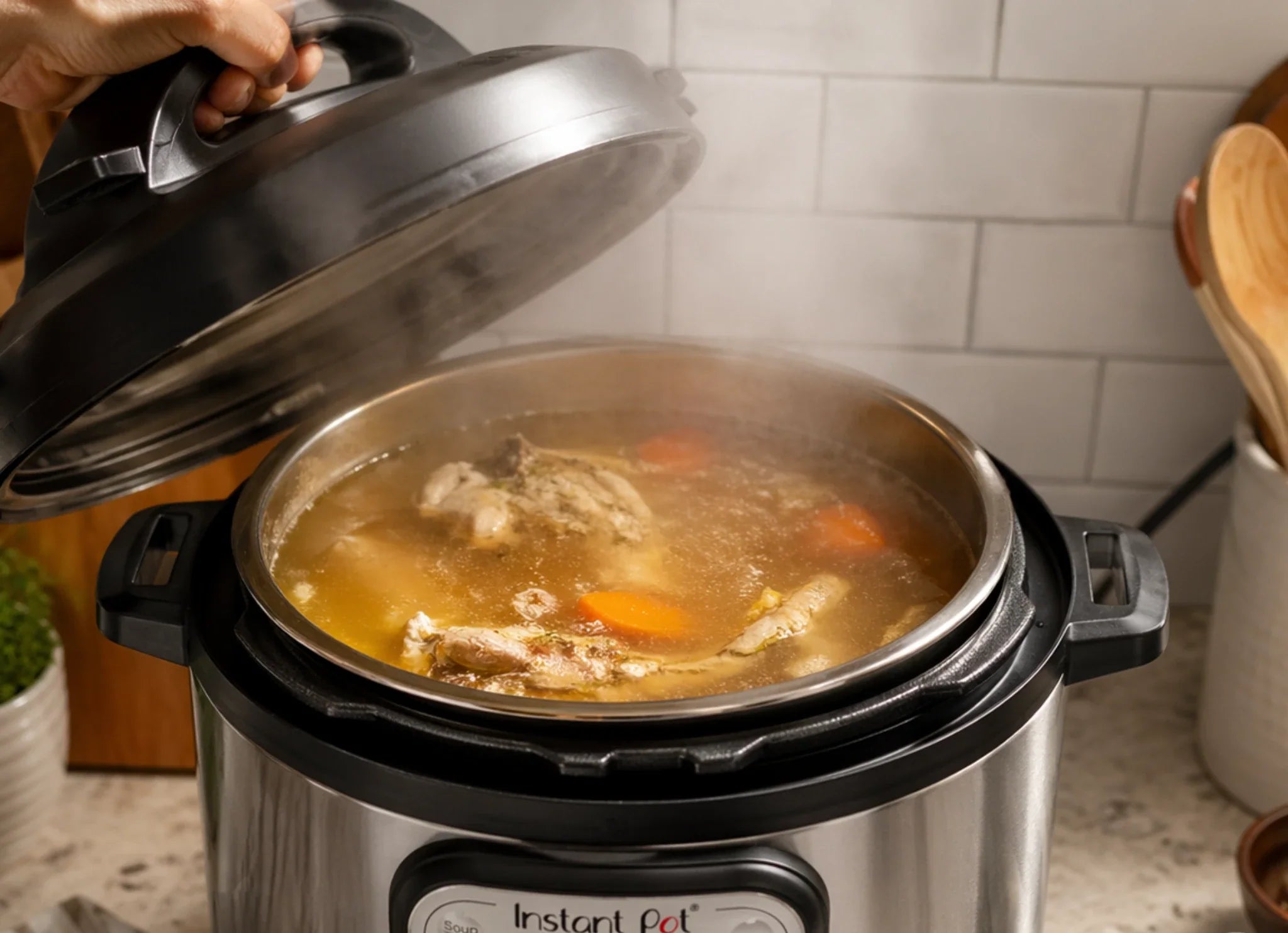Hand lifting Instant Pot lid to reveal simmering chicken bone broth with carrots and chicken bones in a kitchen