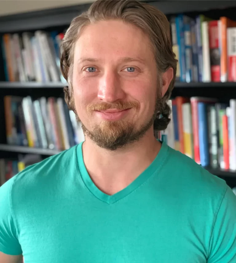 Smiling man with short beard and teal V-neck shirt standing in front of a bookshelf