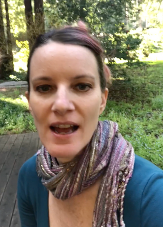 Woman faces the camera outdoors on a wooded path, wearing a blue top and patterned scarf