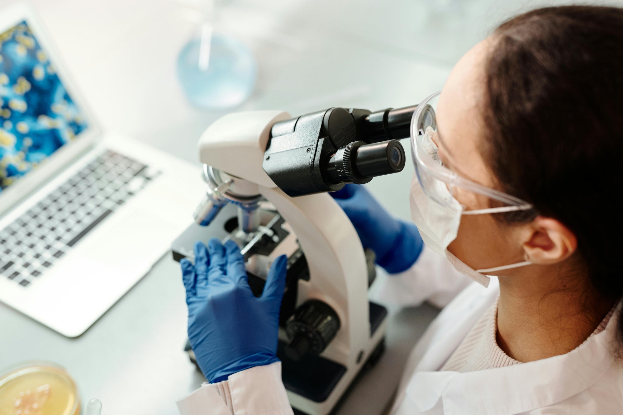 Scientist wearing mask and safety goggles using a microscope, gloved hands adjusting a slide beside an open laptop.