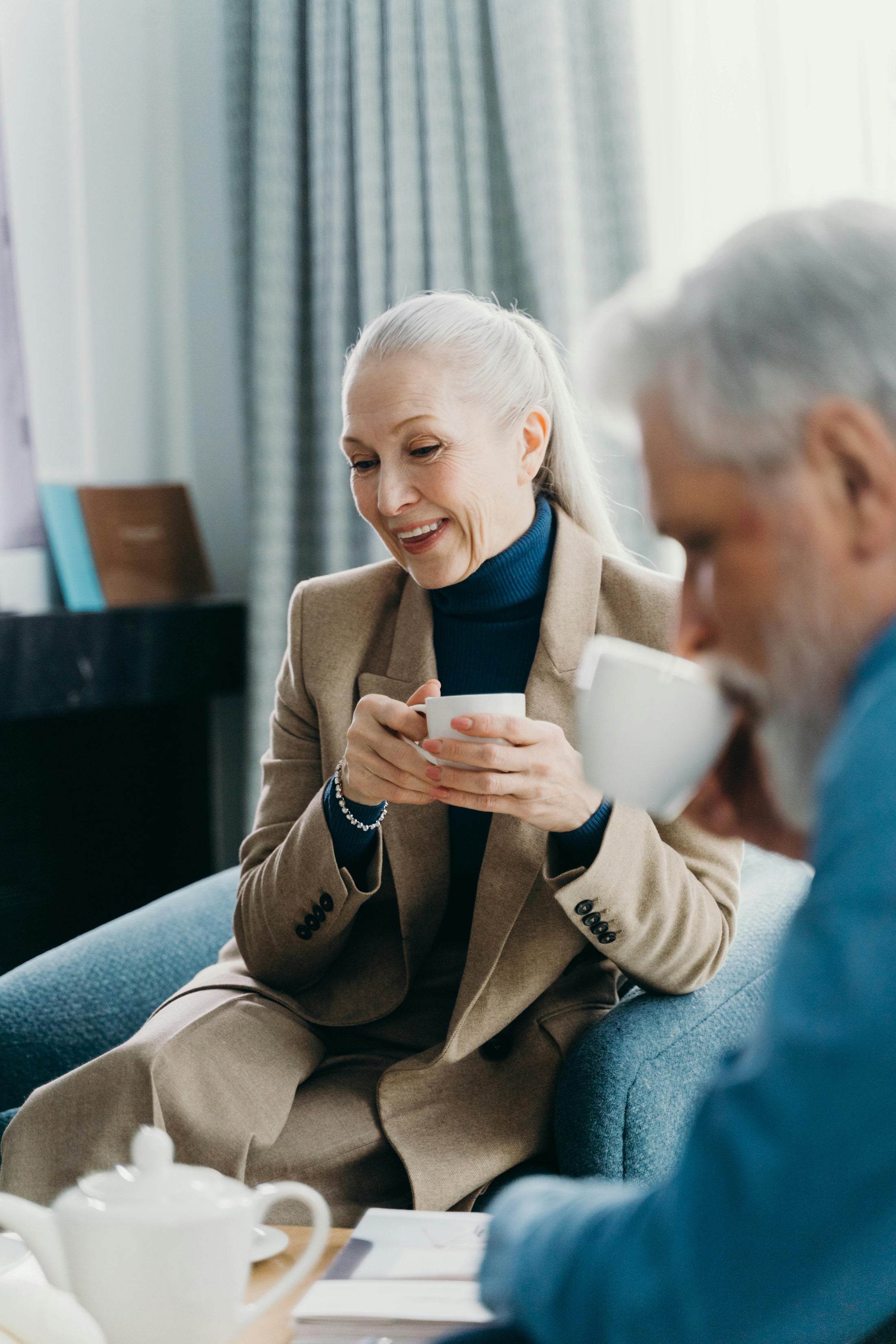 Older woman in tan suit and navy turtleneck smiling while holding a teacup beside a man in conversation