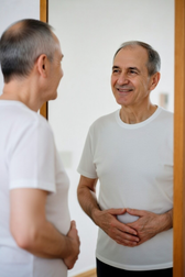 Older man smiling and holding his lower abdomen while looking in a bathroom mirror, implying improved digestion