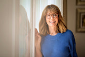 Smiling middle-aged woman with glasses wearing a blue sweater, standing by a window and looking at the camera.