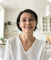 Smiling middle-aged woman with short dark hair wearing a white blouse, seated in a bright home kitchen