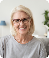 Smiling older woman with short blonde hair and red-framed glasses seated on a living room sofa