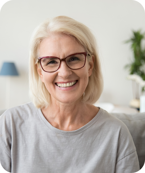 Smiling older woman with short blonde hair and red-framed glasses seated on a living room sofa