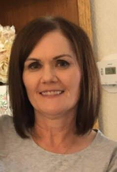 Smiling middle-aged woman, shoulder-length brown hair, gray top; indoor setting with thermostat visible.