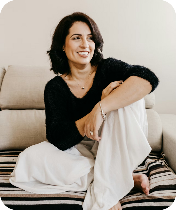 Smiling woman with dark hair in a black sweater and white pants sitting cross-legged on a striped cushion, looking right.