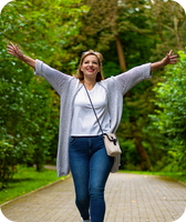 Woman smiling with arms outstretched on tree-lined park path, wearing white shirt, gray cardigan, jeans and crossbody bag.