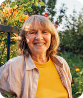Smiling older woman in a yellow shirt and light jacket, standing in a sunny flower garden with roses.