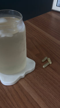 Iced beverage in a glass on a white coaster beside three green supplement capsules on a wooden table.